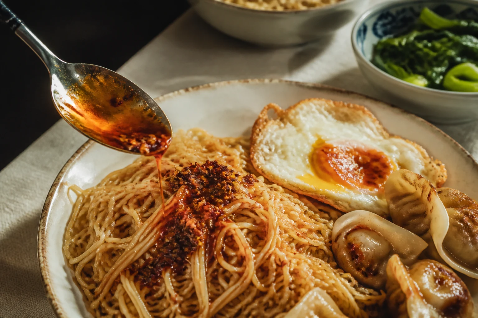 Chilli oil being spooned over noodles with a fried egg and dumplings